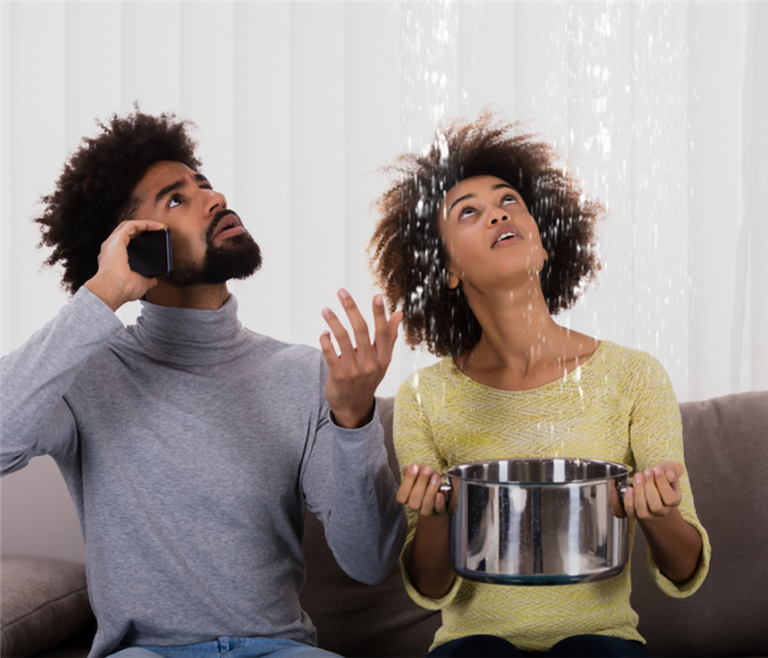 picture shows a man and a woman looking up to a leaky roof while holding a pot/ 